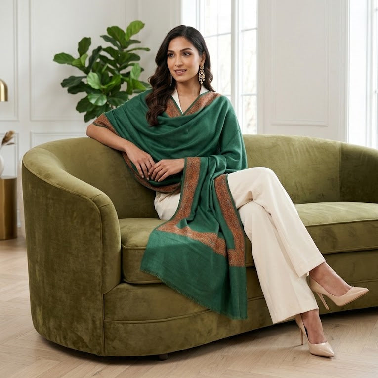 A woman wearing a green shawl draped over her shoulders, cream wide-leg trousers, and nude heels, sitting on an olive green velvet sofa in a bright, modern living room.