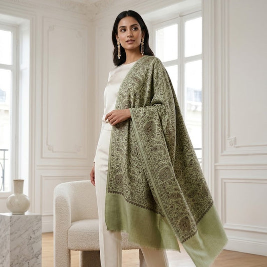 A woman stands in a bright white room with wood floors, wearing a white outfit and holding a large sage green shawl with an intricate dark green border pattern.