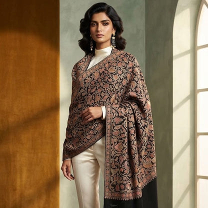 A woman models a brown and black patterned shawl draped over her shoulders, paired with cream trousers and black heels in a softly lit interior with warm wood and textured walls.