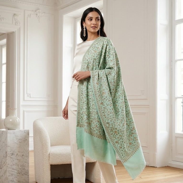 A woman models a light mint green shawl with a delicate floral pattern and a solid border, styled over a white dress in a bright, classic European room with white walls and wooden floors.