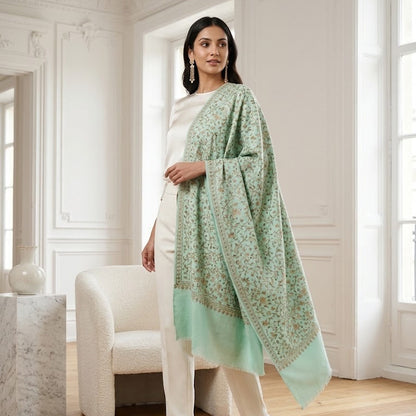 A woman models a light mint green shawl with a delicate floral pattern and a solid border, styled over a white dress in a bright, classic European room with white walls and wooden floors.