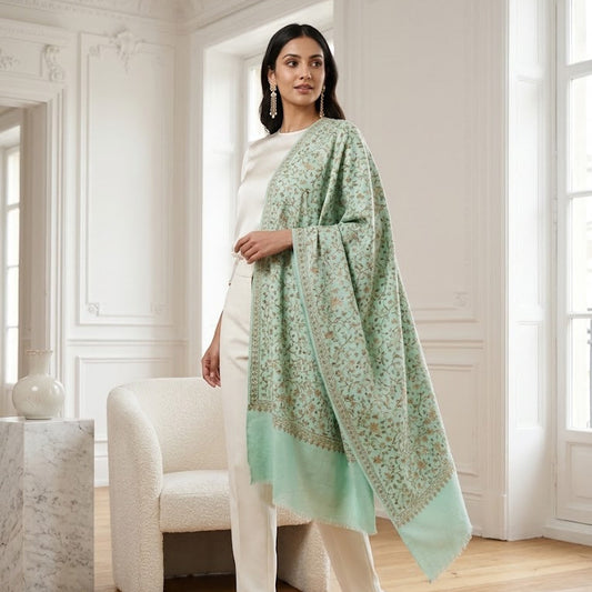 A woman models a light mint green shawl with a delicate floral pattern and a solid border, styled over a white dress in a bright, classic European room with white walls and wooden floors.