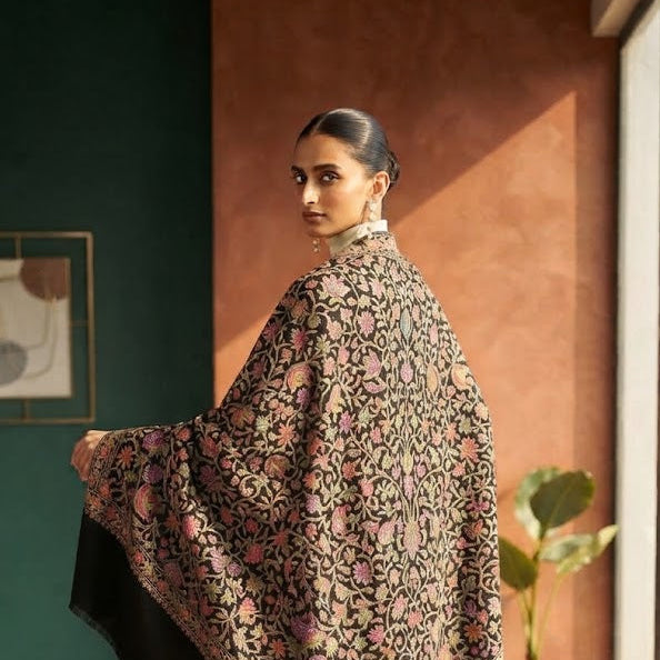A woman wearing a black shawl with an intricate pink and gold floral pattern, paired with white trousers and beige heels, standing in a warmly lit indoor setting.