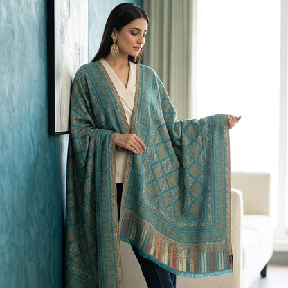 A woman models a teal and gold patterned shawl draped over her shoulders in a bright room with a blue wall and a white armchair.
