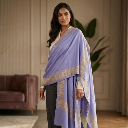 A woman models a lavender purple shawl with gold printed patterns, draped over her shoulders, paired with dark gray wide-leg trousers and beige heels in a warm, elegant room.