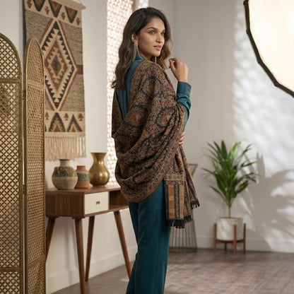 A woman models a dark brown and black patterned shawl draped over her shoulders, paired with teal pants and heels, in a styled indoor room with bohemian decor.