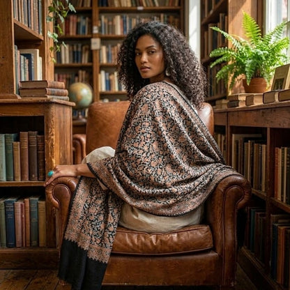 A woman with curly dark hair sits in a brown leather armchair inside a cozy, book-filled library, draped in a patterned shawl with a black border.