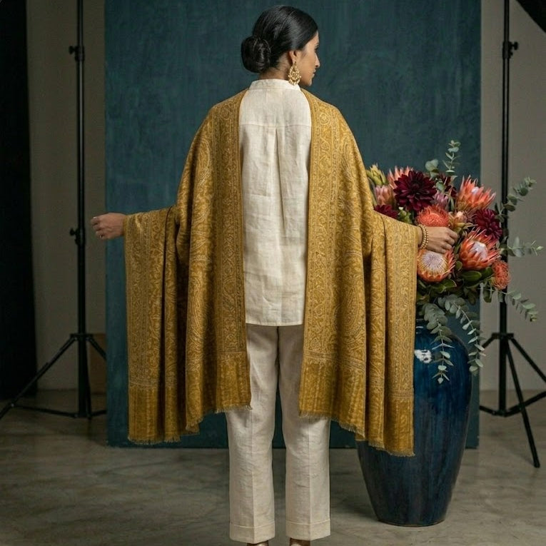 Person wearing a mustard yellow garment with a white outfit, standing in front of a blue backdrop with floral arrangements.