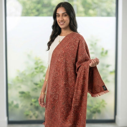 A woman modeling a rust-colored, patterned shawl with fringe trim over a cream top and blue jeans, standing in a bright room with large windows showing green foliage outside.
