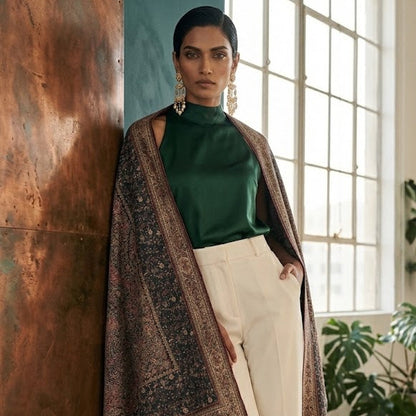 A woman models a dark brown patterned shawl draped over her shoulders, styled with a green mock neck top and cream wide-leg trousers in a modern loft setting with a copper wall and large windows.