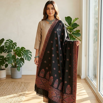 A woman models a black shawl with red and gold traditional patterns, draped over a beige kurta in a bright, modern room with potted plants and large windows.