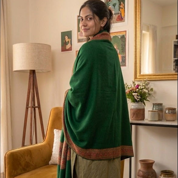 A woman stands in a living room wearing a dark green shawl with a brown border over an olive green outfit. The room features a mustard yellow armchair, a floor lamp, and a patterned rug.
