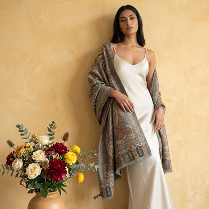 Woman in a white dress holding a patterned shawl next to a vase of flowers against a beige wall.