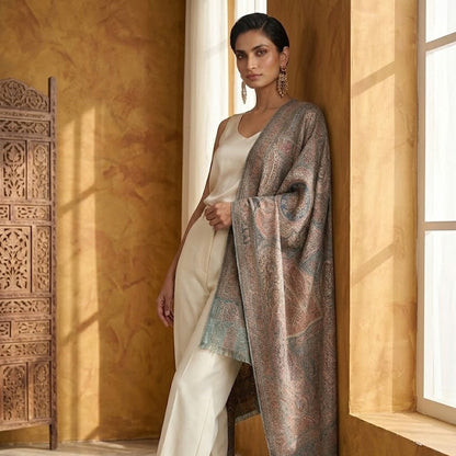 A woman models a traditional outfit featuring a cream-colored kurta and wide-leg trousers, draped with a large, ornate patterned shawl in muted brown, beige, and teal tones, set against a warm, textured yellow wall.