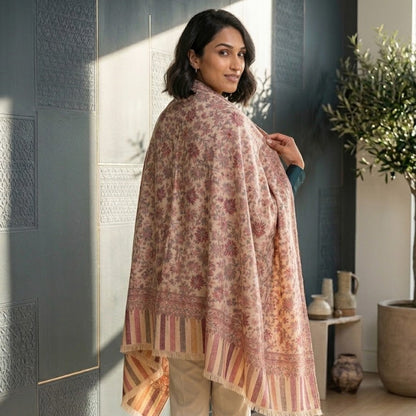 A woman wearing a pink and beige patterned shawl draped over her shoulders, standing in a modern room with a dark gray paneled wall and a potted plant in the background.