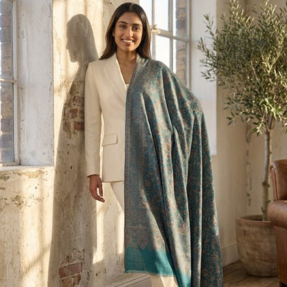 A woman wearing a cream blazer and trousers stands in a sunlit room draped in a large teal and blue patterned shawl with a turquoise gradient hem.