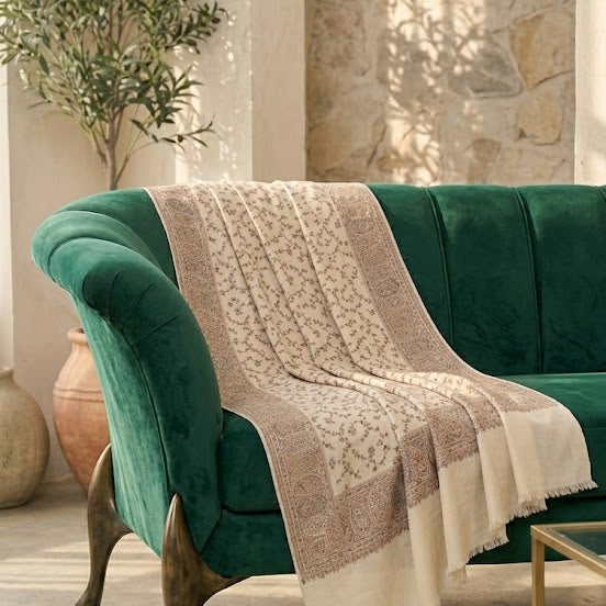 A green velvet armchair in a sunlit room with stone walls and a window, draped with a cream and beige patterned throw blanket.