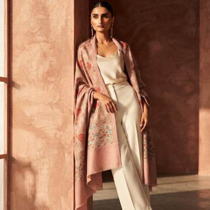 A woman models a light pink shawl with delicate floral embroidery and a white pantsuit in a warm, sunlit room with textured terracotta walls.