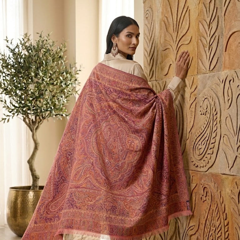 A woman wearing a beige kurta and trousers models a large, draped dusty rose and purple paisley shawl with fringe, standing in a warm, sunlit room next to a carved wooden wall panel.