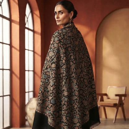 A woman stands in a warm-toned indoor room wearing a large black shawl with an intricate beige floral pattern and solid black borders. The setting features arched windows, warm orange-brown walls, and a wooden chair in the background.
