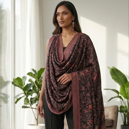 A woman models a dark brown and black patterned shawl draped over her shoulders, paired with black wide-leg trousers, in a bright, modern room with large windows and potted plants.