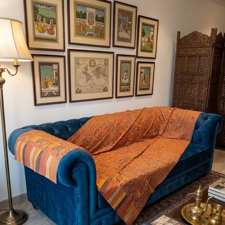 A blue velvet Chesterfield sofa in a styled living room, draped with a vibrant orange and red patterned silk throw blanket. The room features a blue patterned rug, a wooden coffee table with books, a brass floor lamp, and a dark carved wooden screen, with a gallery wall of framed art behind the sofa.