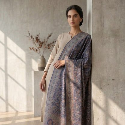 A woman models a traditional draped outfit featuring a sheer, patterned dupatta in muted blue and beige tones over a light beige kurta, set against a minimalist concrete wall with soft window shadows.