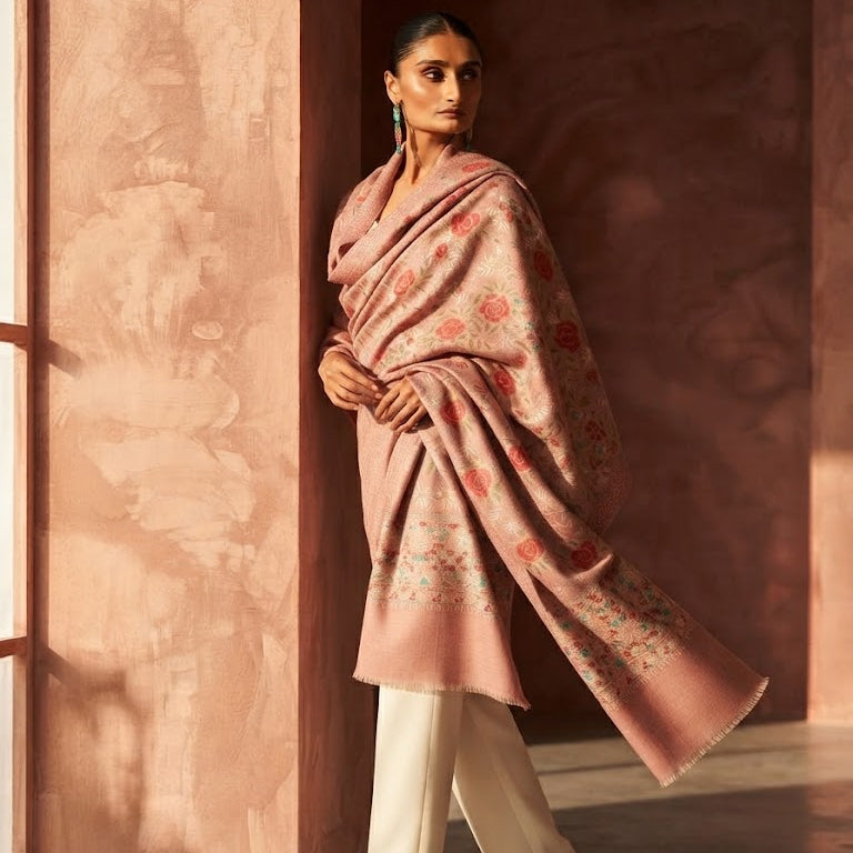 A woman models a dusty pink shawl with delicate floral embroidery, draped over her shoulders and paired with cream trousers, standing in a warm, sunlit room with textured terracotta walls.