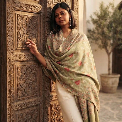 A woman stands beside an ornate carved wooden door wearing a light sage green shawl with red floral motifs and cream trousers, in a sunlit courtyard setting.