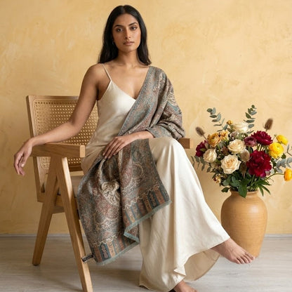 Woman sitting on a wooden chair with a patterned shawl draped over her, against a yellow wall with a vase of flowers.