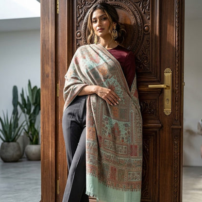A woman stands in front of an ornate wooden door wearing a long, patterned shawl draped over her shoulders, paired with dark trousers, a burgundy top, and dark green heels.