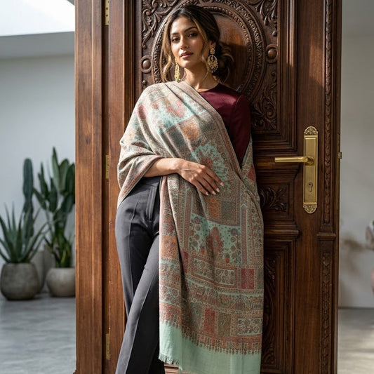 A woman stands in front of an ornate wooden door wearing a long, patterned shawl draped over her shoulders, paired with dark trousers, a burgundy top, and dark green heels.