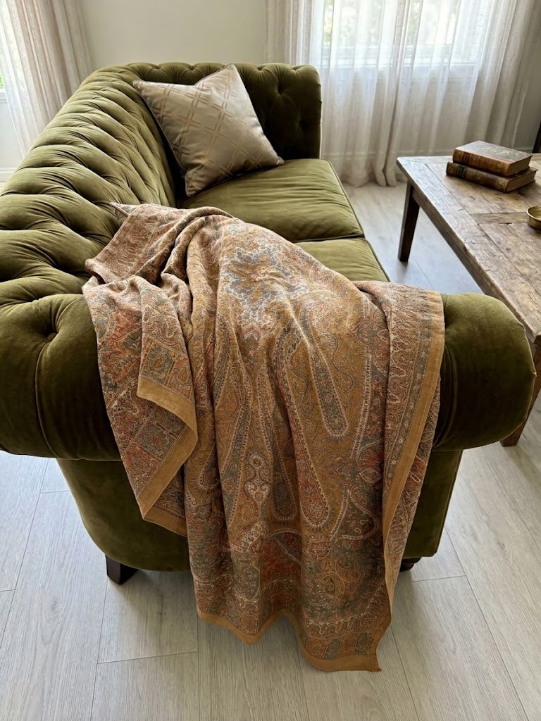 An olive green velvet tufted sofa in a bright living room, draped with a warm beige and rust paisley patterned throw blanket. A matching beige pillow rests on the sofa, with a rustic wooden coffee table and light wood floors visible in the background.