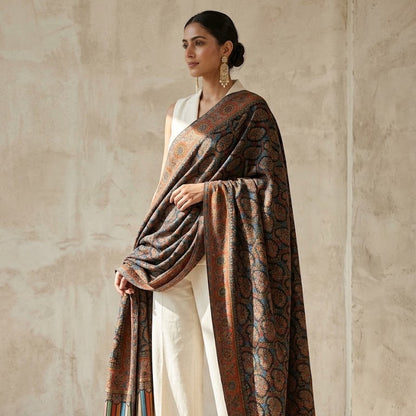 A woman models a luxurious patterned shawl draped over her shoulders, paired with a white sleeveless top and wide-leg trousers. The shawl features an intricate paisley and floral design in brown, rust, and black, with a striped border in red, green, and blue.