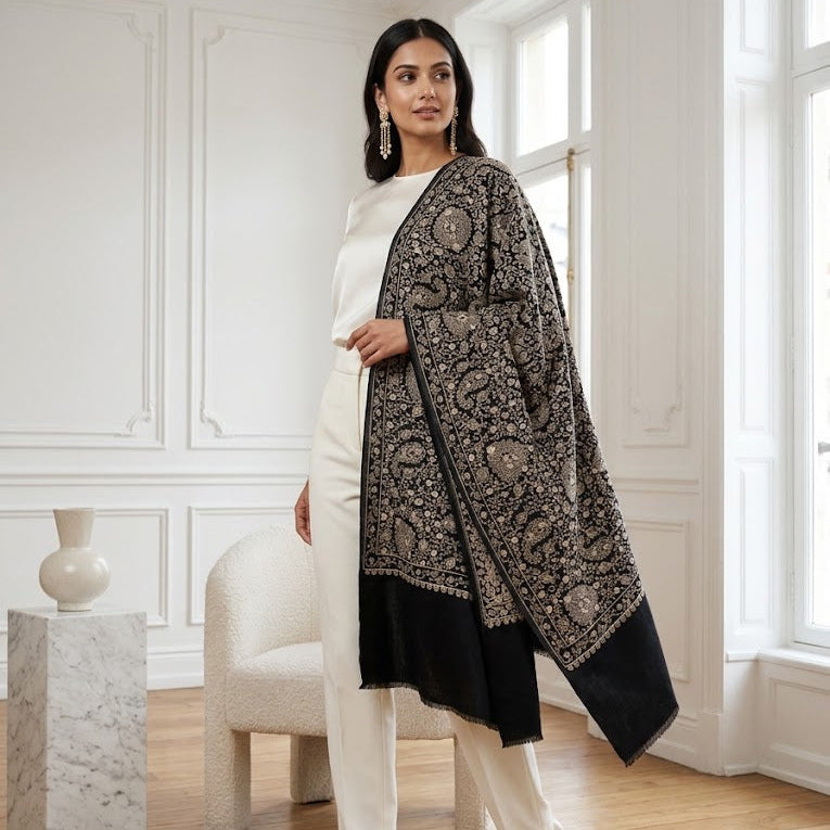 A woman models a black and beige patterned shawl draped over her shoulders, paired with a white top and white trousers in a bright, classic white room with wooden floors.