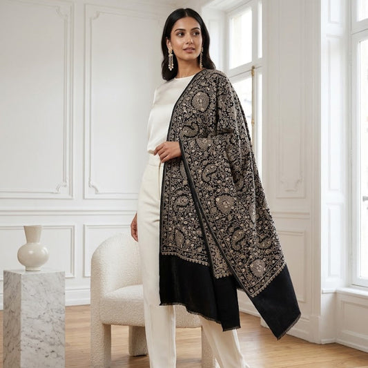 A woman models a black and beige patterned shawl draped over her shoulders, paired with a white top and white trousers in a bright, classic white room with wooden floors.