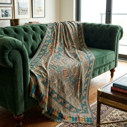 A patterned throw blanket with teal, beige, and brown tones is draped over a green velvet tufted sofa in a styled living room setting.