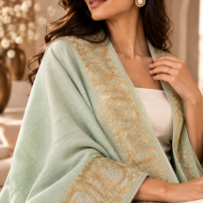 A woman wearing a pale mint green shawl with ornate gold paisley embroidery, draped over her shoulders and arms.