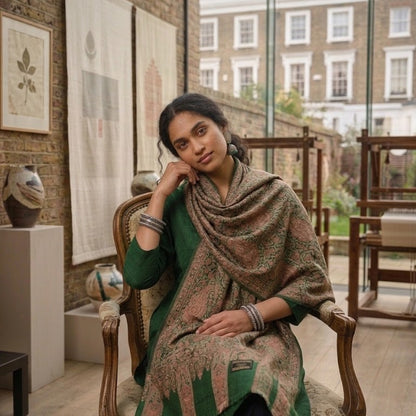 A woman sits in an ornate wooden chair inside a gallery or studio space, wearing a green kurta with a large beige and brown patterned shawl draped over her shoulders. The background features brick walls, framed botanical prints, and large windows looking out onto a brick building.