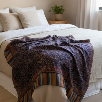 Bed with a patterned blanket draped over the foot, in a room with a plant and framed picture on the wall.
