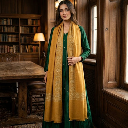 A woman stands in a rustic wooden library wearing a dark green traditional dress with a wide mustard yellow embroidered scarf draped over her shoulders.