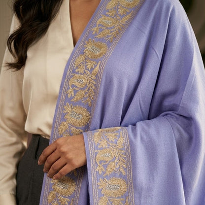 A close-up of a person wearing a lavender blue shawl with ornate gold floral embroidery along the borders, paired with a cream blouse and dark trousers.