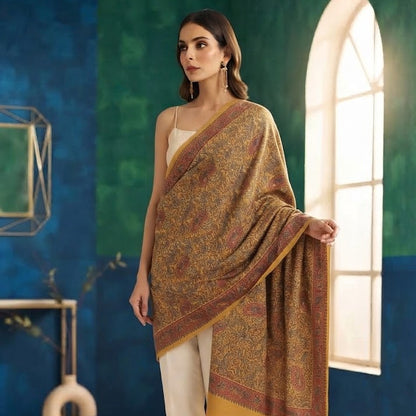 A woman models a traditional draped shawl or scarf in a warm brown and gold paisley pattern, styled over cream pants in an indoor setting with blue and green textured walls and a bright arched window.