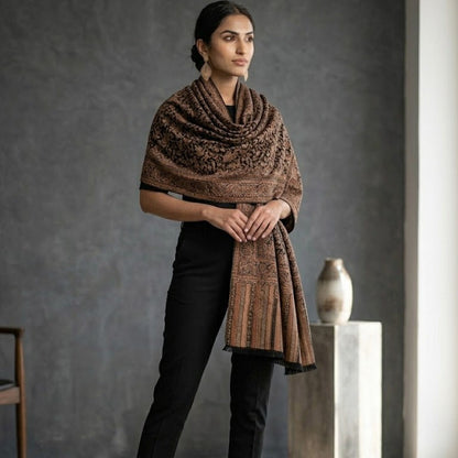 A woman models a brown patterned shawl draped over her shoulders, paired with black pants and beige heels, standing in a modern studio with a textured gray wall.