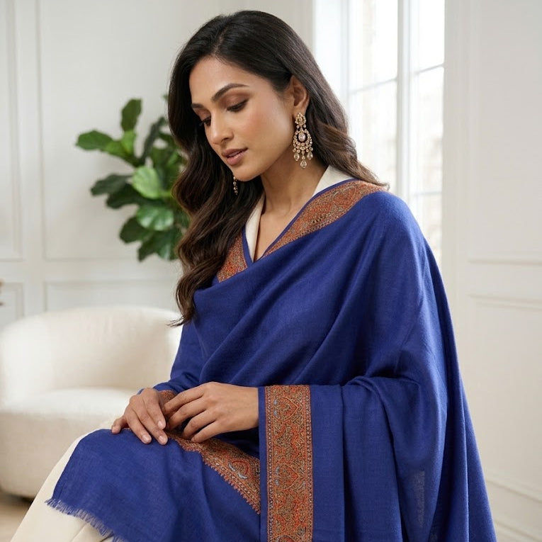 A woman seated indoors wearing a royal blue shawl with ornate brown border detailing and cream trousers. The setting is a bright, modern room with a white sofa and a green plant in the background.