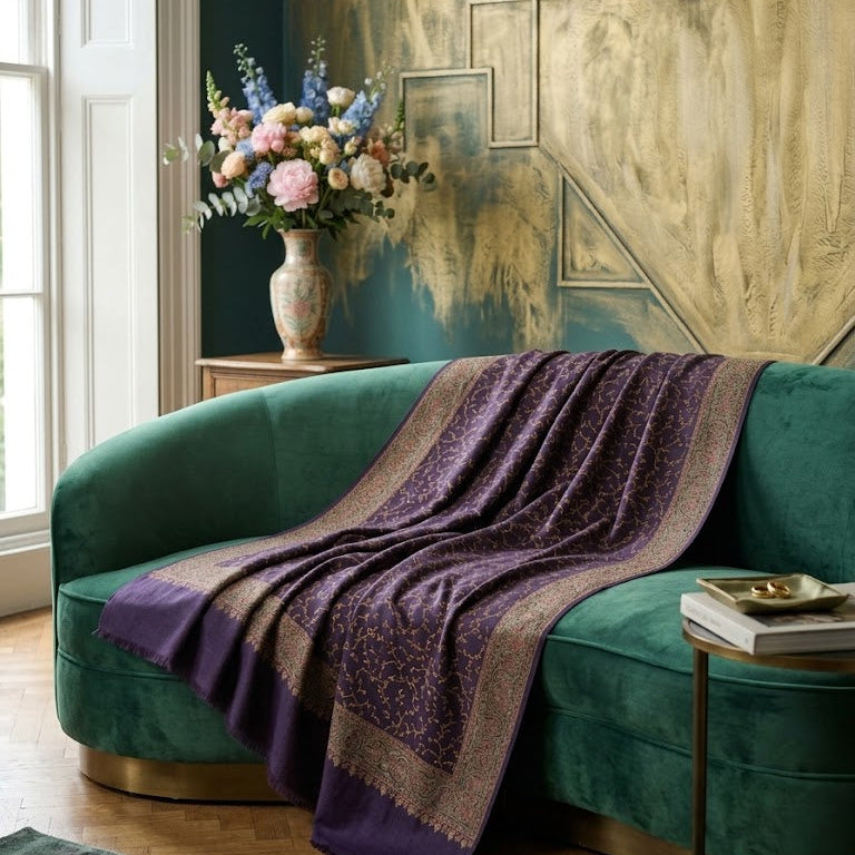 A purple patterned shawl with beige borders draped over a curved emerald green velvet sofa in a styled living room setting.