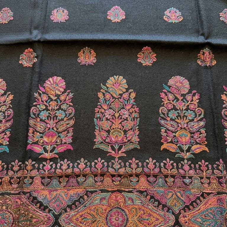 A close-up of a dark charcoal gray fabric featuring vibrant, multicolored floral and geometric embroidery. The fabric displays a repeating pattern of small pink and orange floral motifs at the top, three large central floral motifs in pink, orange, and green, and a wide decorative border at the bottom with intricate paisley and mandala designs.