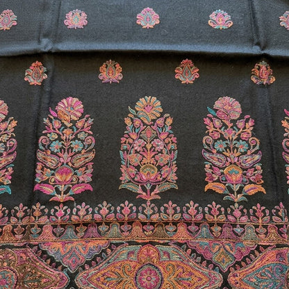 A close-up of a dark charcoal gray fabric featuring vibrant, multicolored floral and geometric embroidery. The fabric displays a repeating pattern of small pink and orange floral motifs at the top, three large central floral motifs in pink, orange, and green, and a wide decorative border at the bottom with intricate paisley and mandala designs.