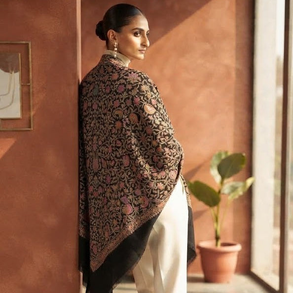 A woman stands in a sunlit indoor corridor wearing a dark patterned shawl draped over her shoulders, cream trousers, and nude heels. The background features warm terracotta walls and a potted plant.