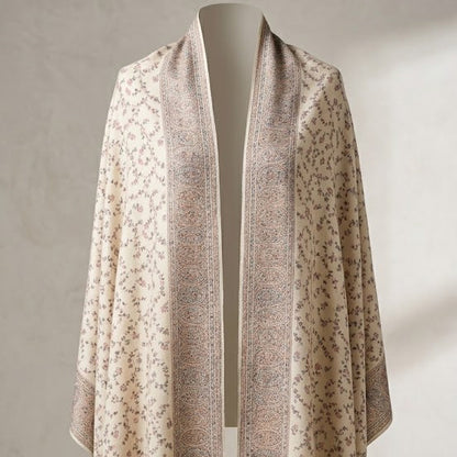A lightweight open-front shawl draped over a mannequin, featuring a cream base with delicate floral motifs and wide taupe-brown patterned borders, set against a neutral textured wall.